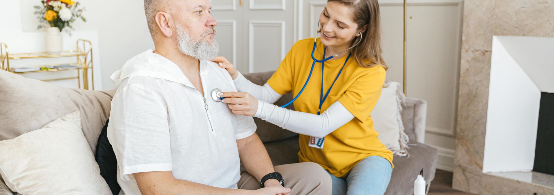A nurse uses a stethoscope for a home check-up on a senior adult in a cozy living room.