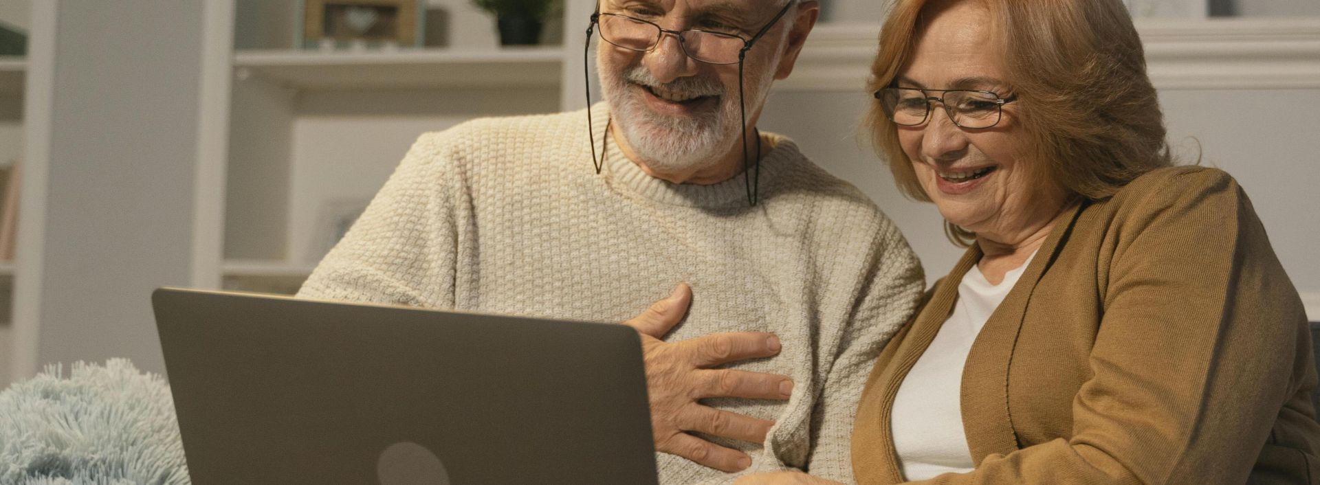 Happy senior couple sitting on a sofa having a joyful video call on a laptop in a cozy living room.